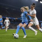 Suasana pertandingan sepak bola wanita di stadion pada malam hari dengan dua tim berseragam biru muda dan putih beradu di lapangan.