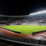 Lapangan stadion sepak bola Indonesia yang diterangi lampu untuk pertandingan malam dengan tribun dan area operasional yang tertata